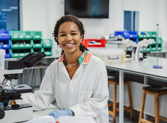 Rhea Fofana smiling whilst in laboratory setting. She is sat a workbench in full lab gear and brightfield microscope is ready to view a slide of unknown contents.